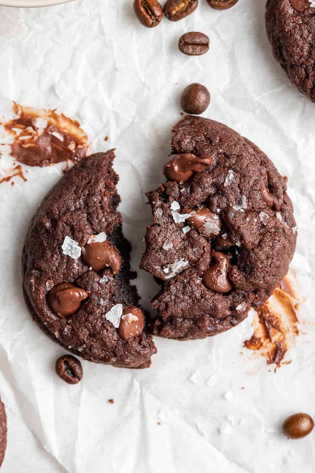 An espresso cookie broken in half, with chocolate smeared on the table, surrounded by espresso beans