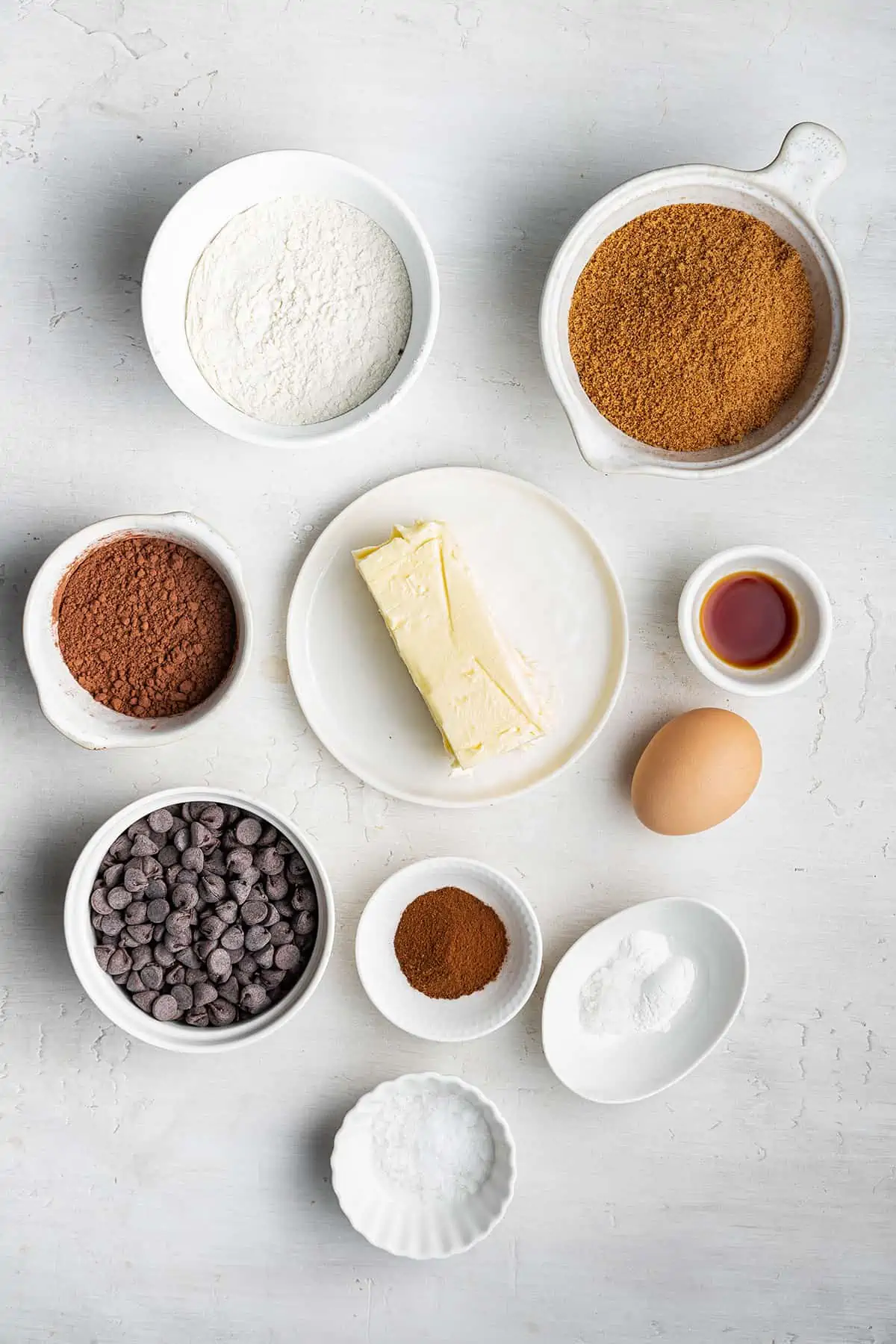 Overhead view of the ingredients for espresso cookies: flour, cocoa powder, espresso powder, chocolate chips, vegan butter, baking powder, baking soda, salt, vanilla extract, and an egg