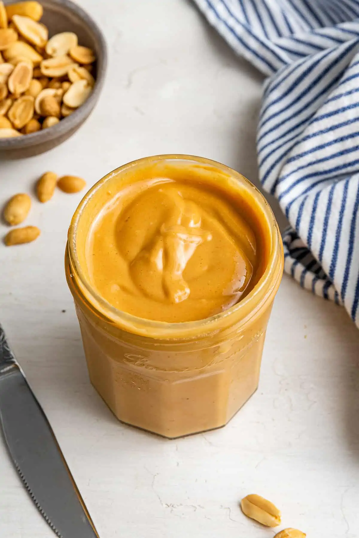 A jar of honey roasted peanut butter, with peanuts and a kitchen towel in the background
