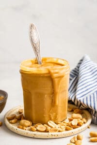 A jar of honey roasted peanut butter on a plate, with a spoon sticking out of it, peanuts on the plate, and a blue and white striped kitchen towel