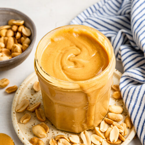 A jar of honey roasted peanut butter on a plate covered with peanuts, next to a bowl of peanuts and a kitchen towel