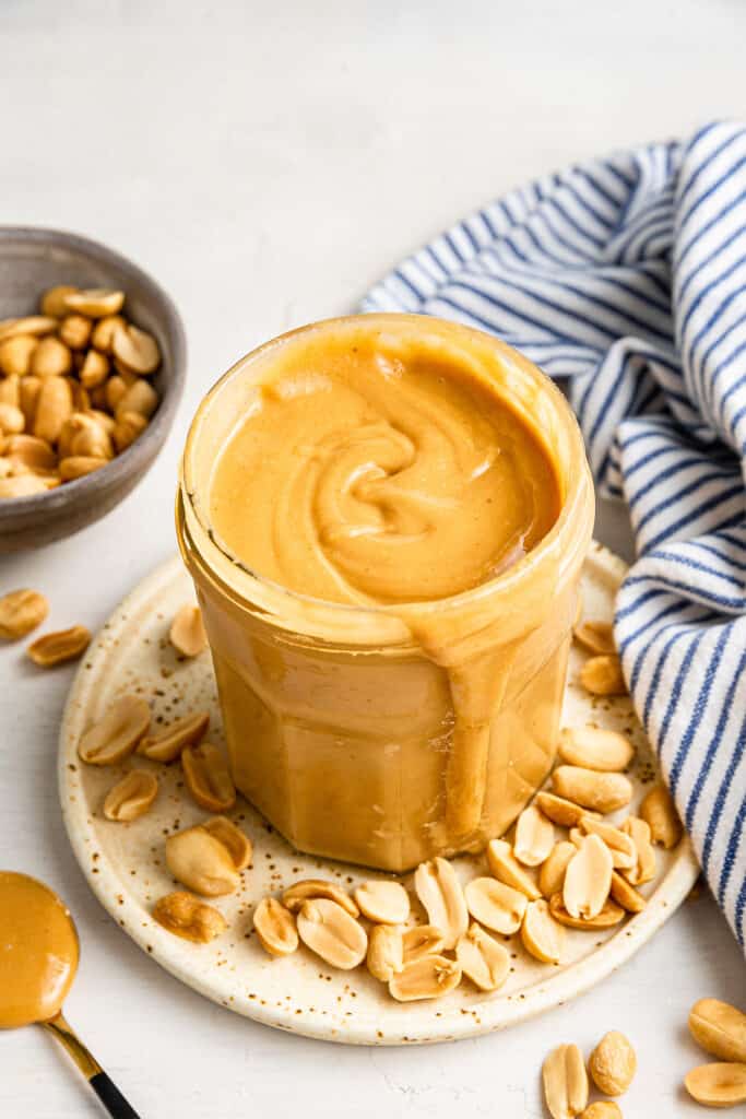 A jar of honey roasted peanut butter on a plate covered with peanuts, next to a bowl of peanuts and a kitchen towel
