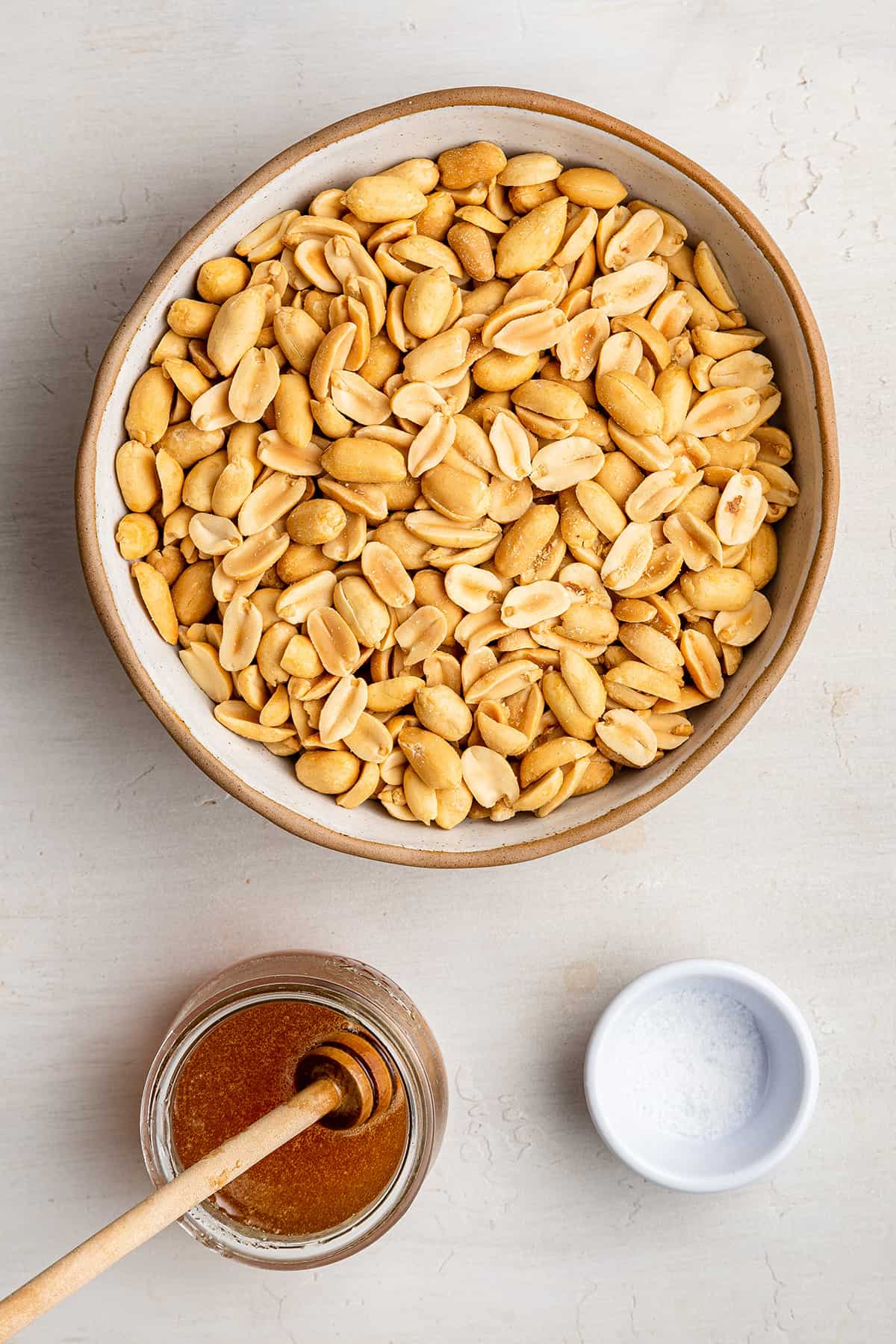 Visão aérea dos ingredientes necessários para a manteiga de amendoim torrada com mel: uma tigela de amendoim, uma jarra de mel com um agitador de madeira e uma tigela de sal