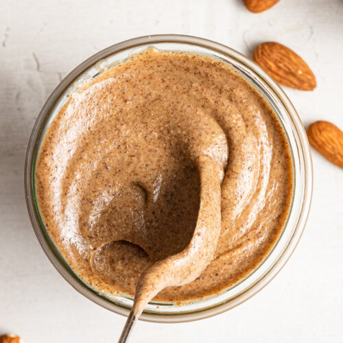 Overhead view of a jar of pumpkin spice almond butter with a spoon lifting out of it, surrounded by almonds