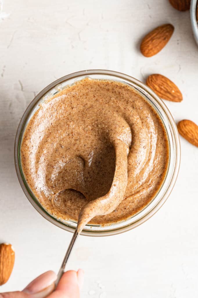 Overhead view of a jar of pumpkin spice almond butter with a spoon lifting out of it, surrounded by almonds