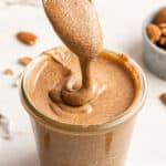 A spoonful of pumpkin spice almond butter drizzling the almond butter into a full jar of it, with a bowl of almonds in the background