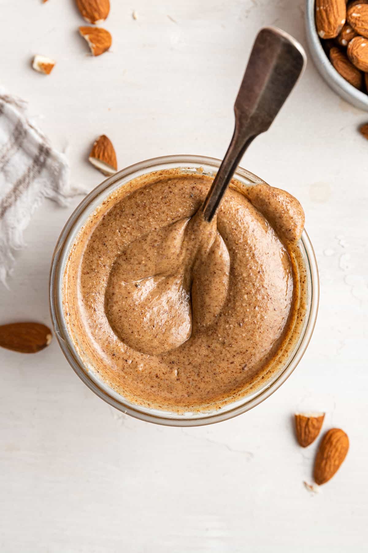 Overhead view of a jar of pumpkin spice almond butter, with a spoon fully submerged in it, surrounded by almonds and a kitchen towel