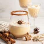 A glass of Southern Comfort eggnog, topped with whipped cream and a star anise pod, with two glasses of eggnog in the background, and cinnamon sticks in the foreground, with the glass next to a pinecone