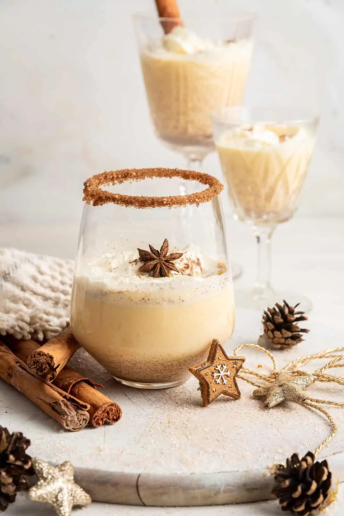 A glass of Southern Comfort eggnog, topped with whipped cream and a star anise pod, with two glasses of eggnog in the background, and cinnamon sticks in the foreground, with the glass next to a pinecone