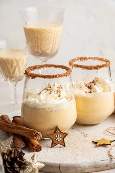 Two glasses of Southern Comfort eggnog, topped with whipped cream, in rimmed glasses, with cinnamon sticks surrounding them, and two wine glasses with Southern Comfort eggnog in the background