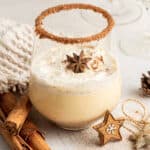 A glass of Southern Comfort eggnog, topped with whipped cream and a star anise pod, with a flavored rim, surrounded bycinnamon sticks