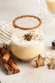 A glass of Southern Comfort eggnog, topped with whipped cream and a star anise pod, with a flavored rim, surrounded bycinnamon sticks