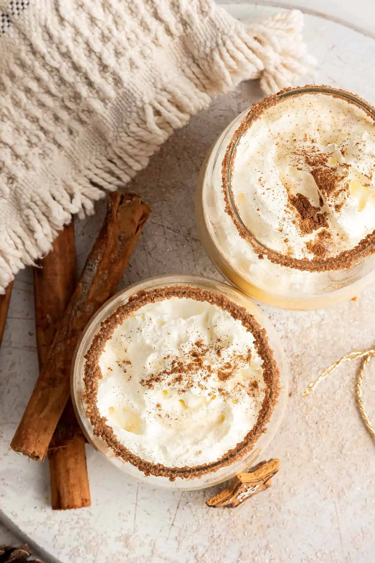 Overhead view of two glasses of Southern Comfort eggnog, topped with whipped cream and ground cinnamon, with a flavored rim, surrounded by cinnamon sticks