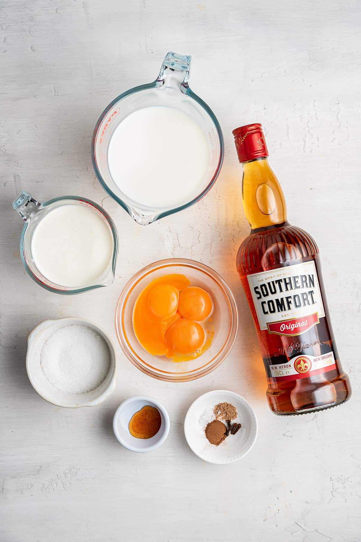Overhead view of a bottle of Southern Comfort, a bowl of egg yolks, a bowl of milk, a bowl of heavy cream, a bowl of sugar, a bowl of vanilla extract, and a bowl of spices