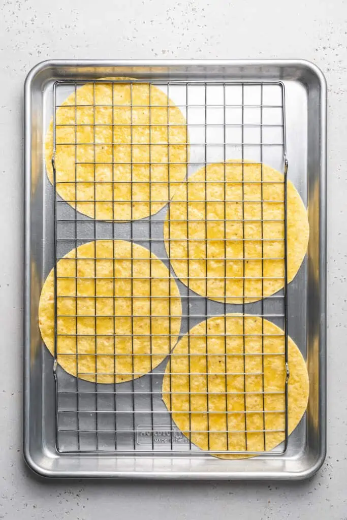 Learn to make homemade tostada shells that are golden-baked, crispy, and seasoned with flaky salt. No frying, and they're ready in 10 minutes! A wire rack covering four corn tortillas on a metal baking sheet.
