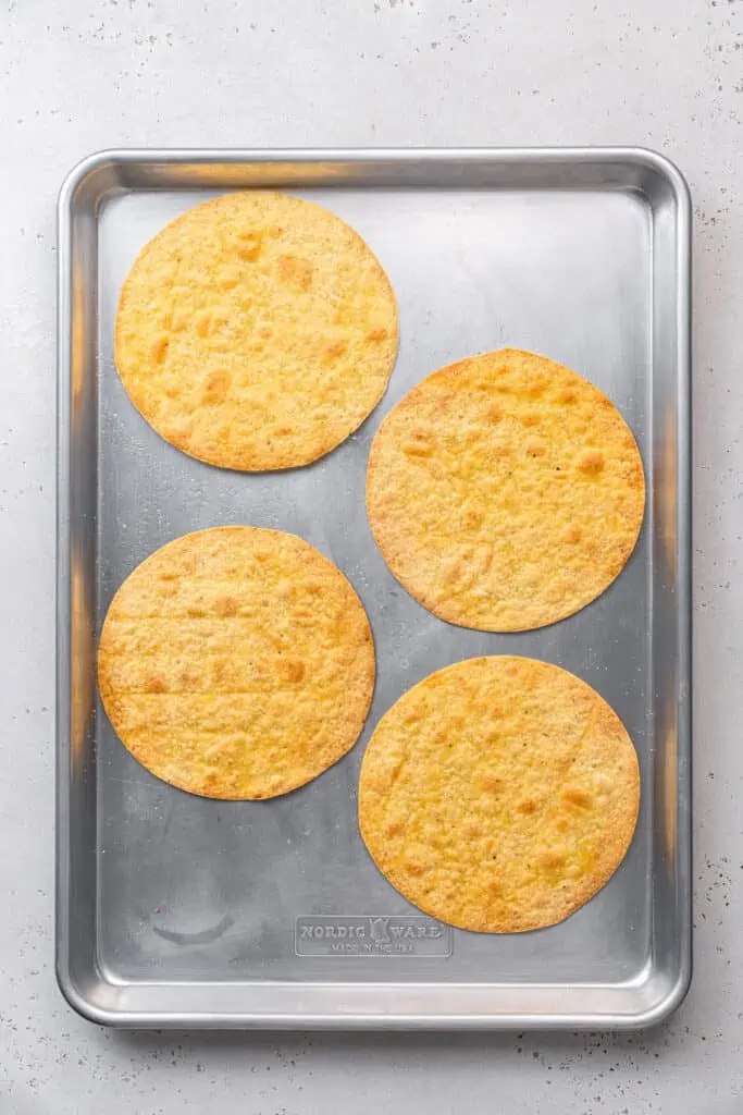 Learn to make homemade tostada shells that are golden-baked, crispy, and seasoned with flaky salt. No frying, and they're ready in 10 minutes! Four baked tostada shells on a metal baking sheet.