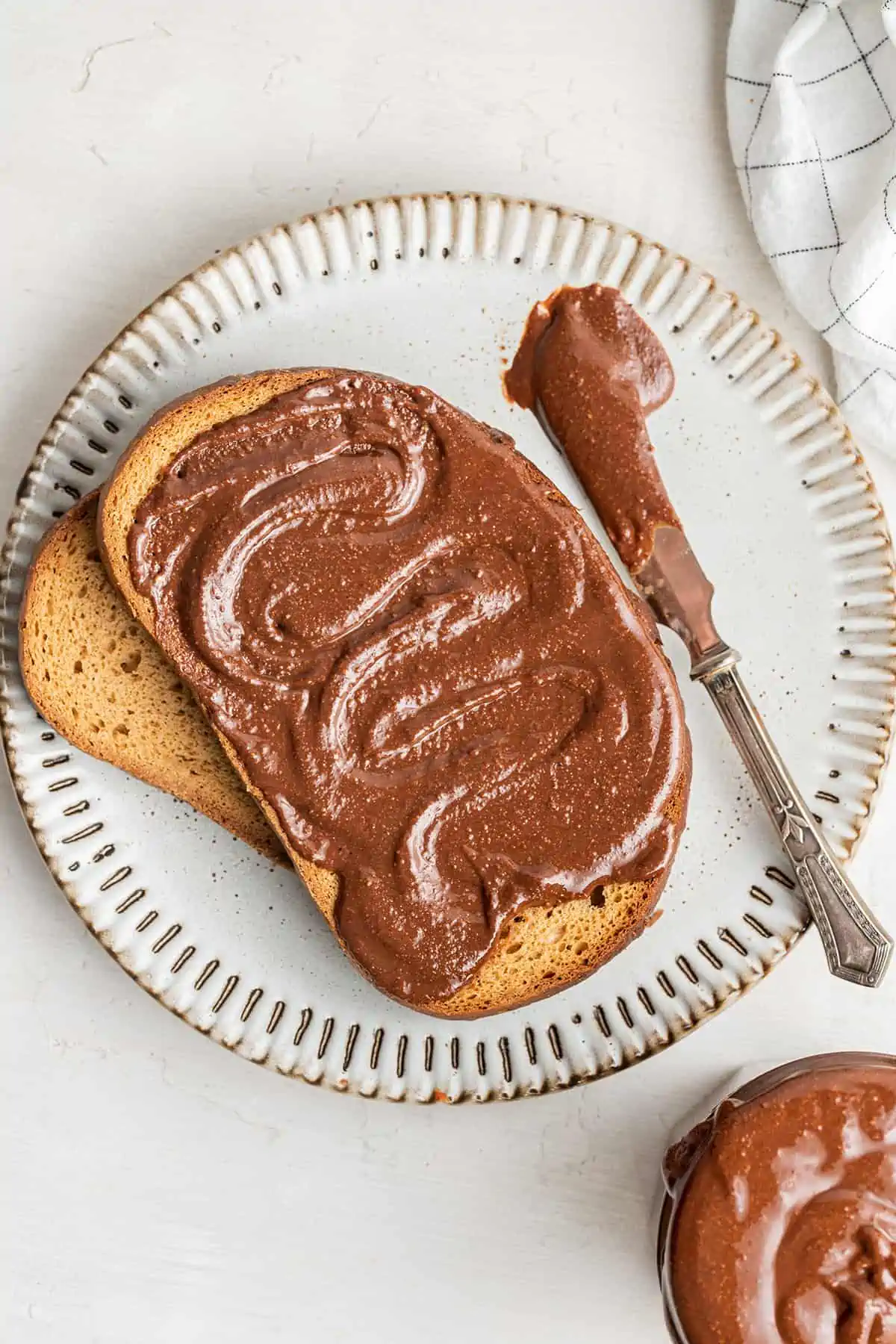 A plate with a piece of toast covered in nutella, on top of another piece of toast and next to a knife covered in nutella