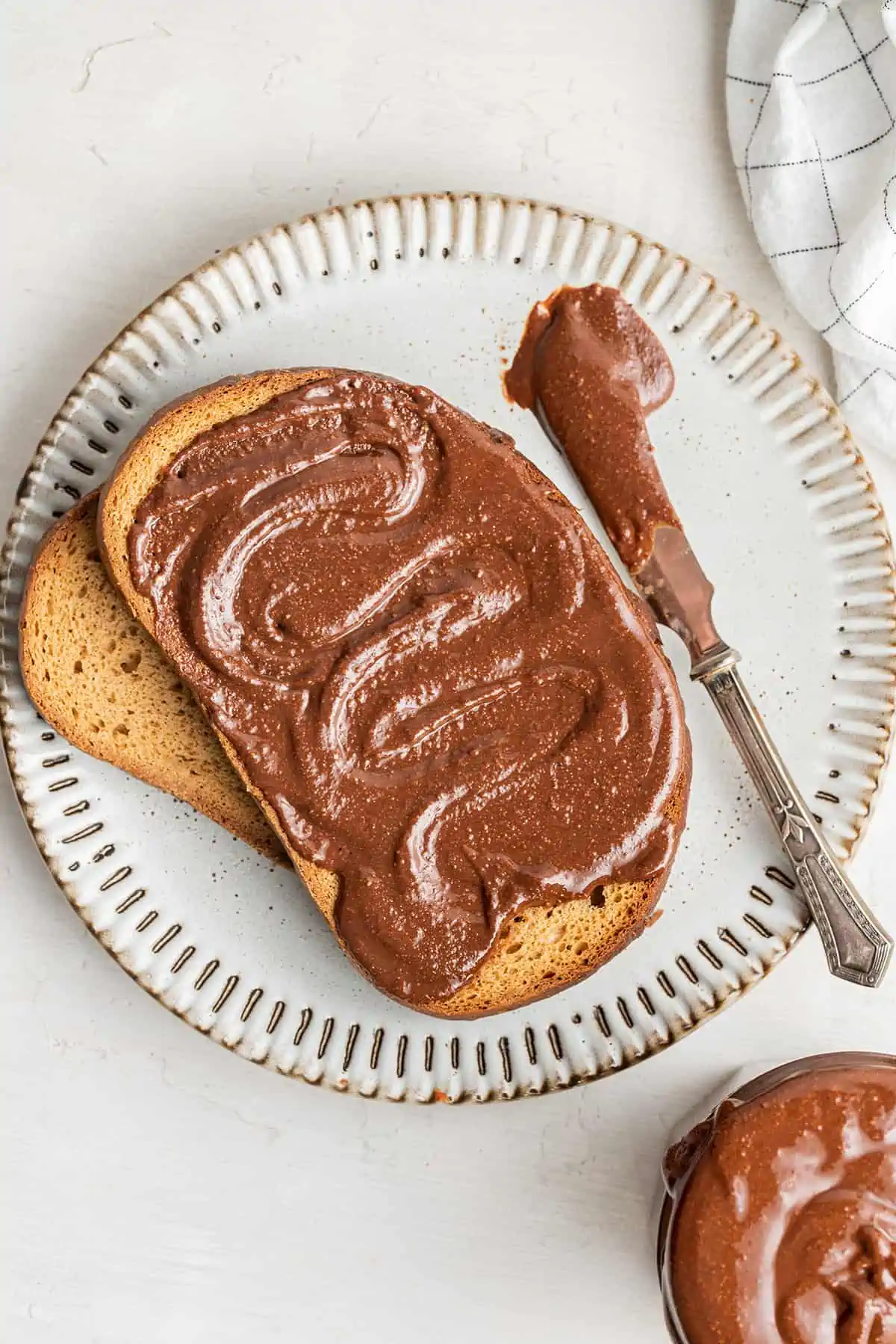 Overhead view of a plate with a piece of toast covered in nutella, on top of another piece of toast and next to a knife covered in nutella.