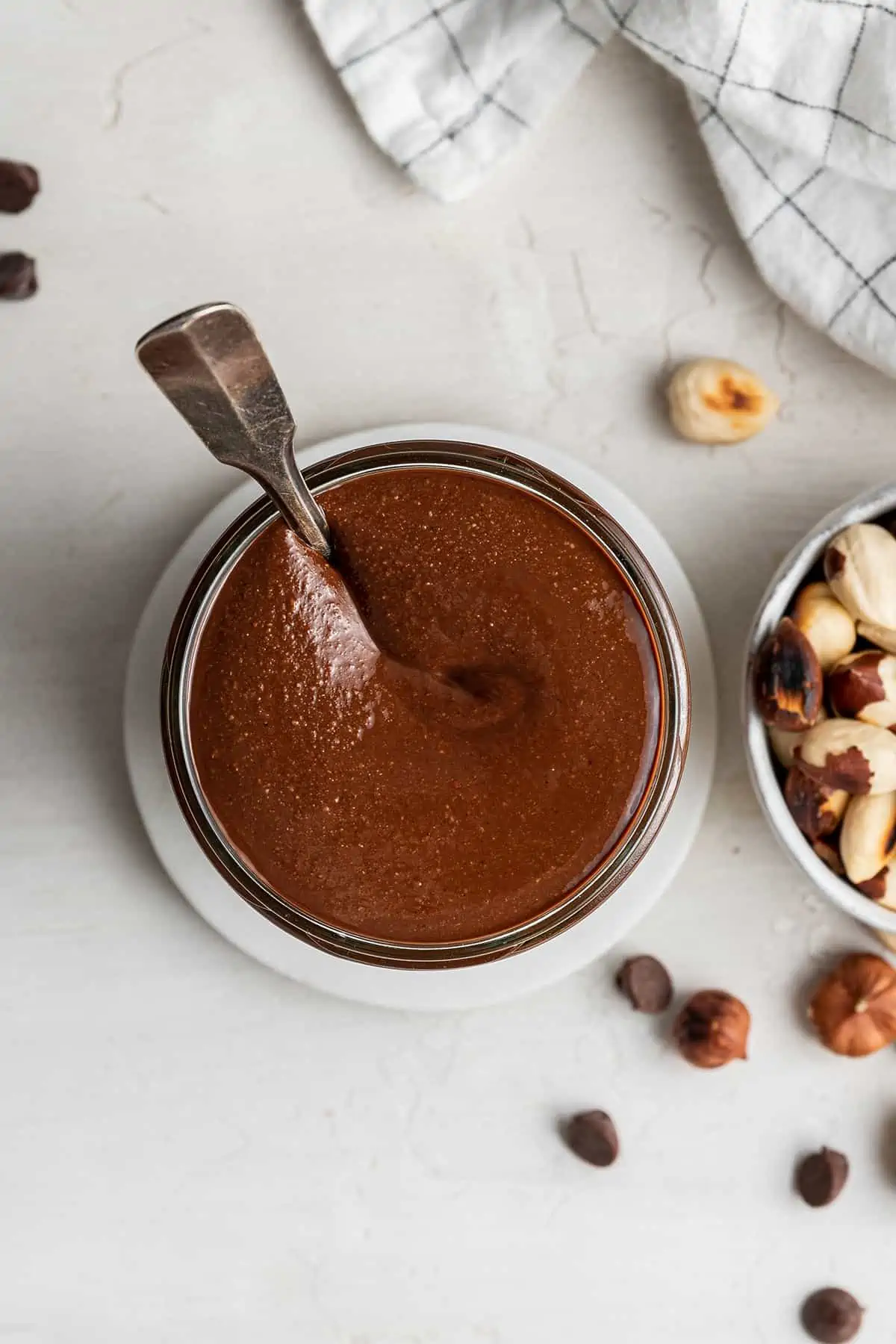 Overhead view of a jar of homemade nutella with a spoon in it, next to some hazelnuts.