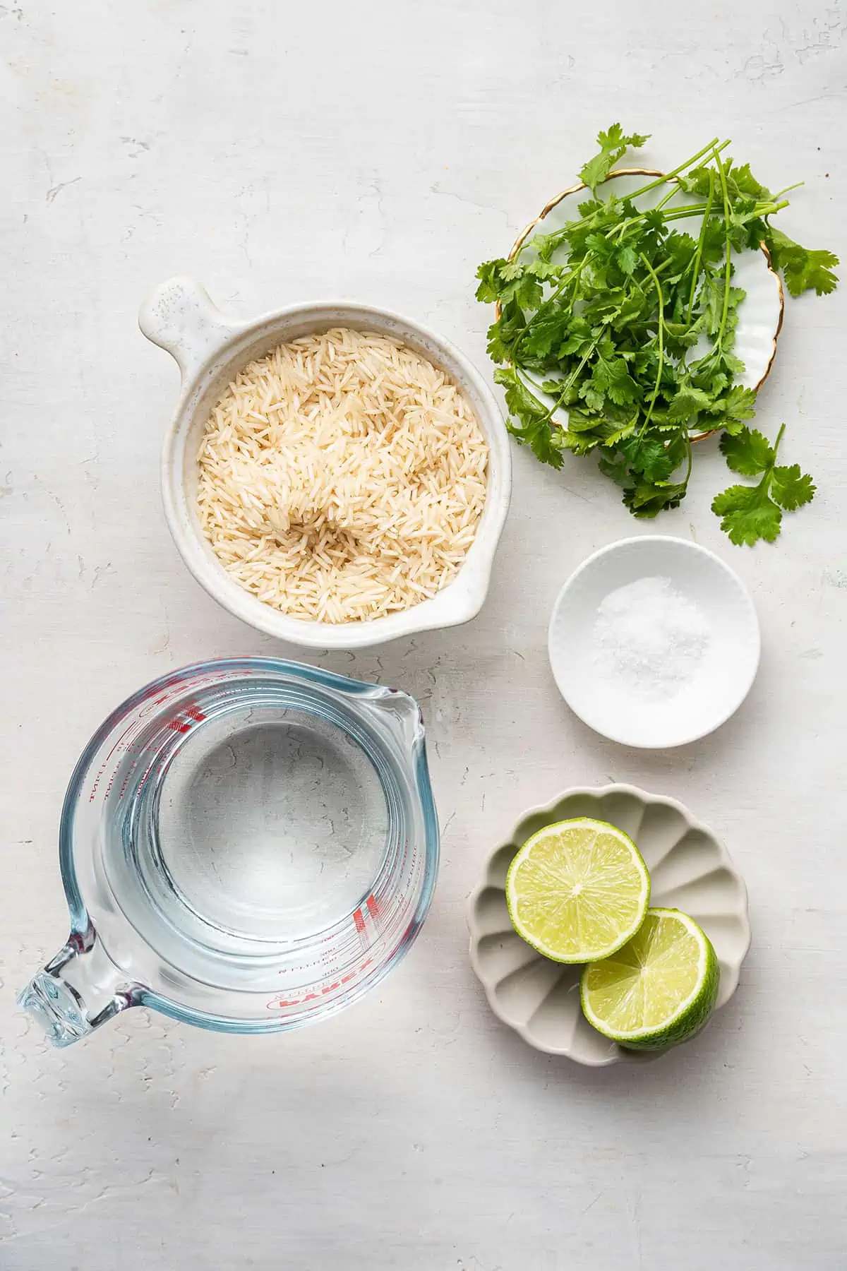 Overhead view of the ingredients for cilantro lime rice: a bowl of raw rice, a bunch of cilantro, a bowl of salt, a bowl with two lime halves, and a pyrex of water.