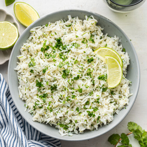 A plate of cilantro lime rice topped with lime slices, next to a sprig of cilantro, fresh limes, a kitchen towel, and a water glass