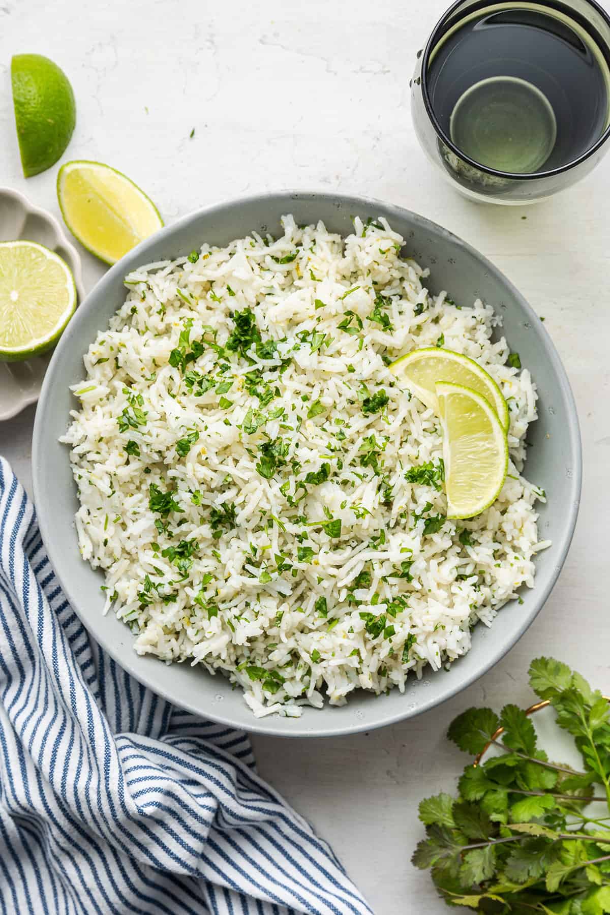 A plate of cilantro lime rice topped with lime slices, next to a sprig of cilantro, fresh limes, a kitchen towel, and a water glass