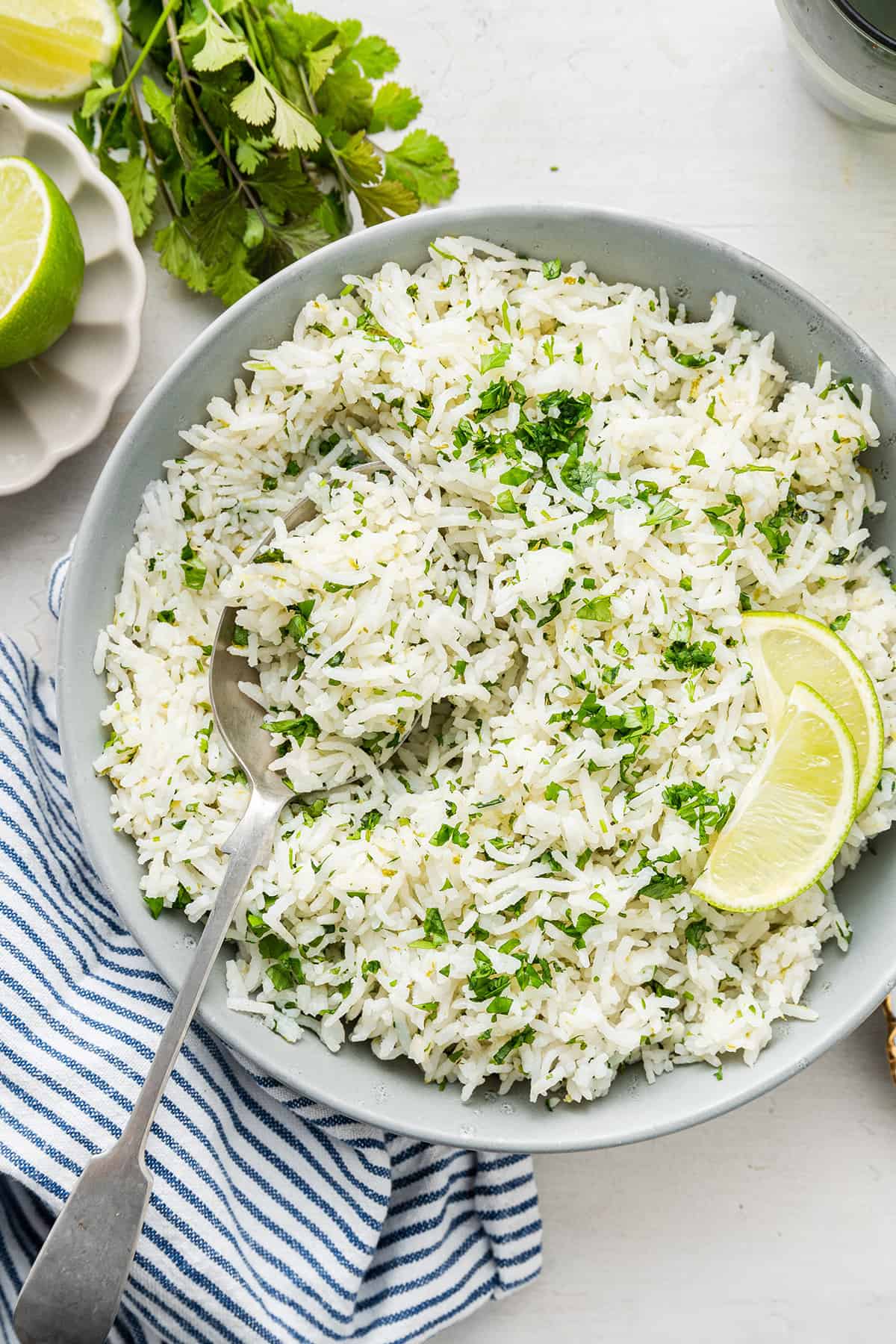 A plate full of cilantro lime rice with a serving spoon in it, and two lime slices, with cilantro and fresh lime next to it