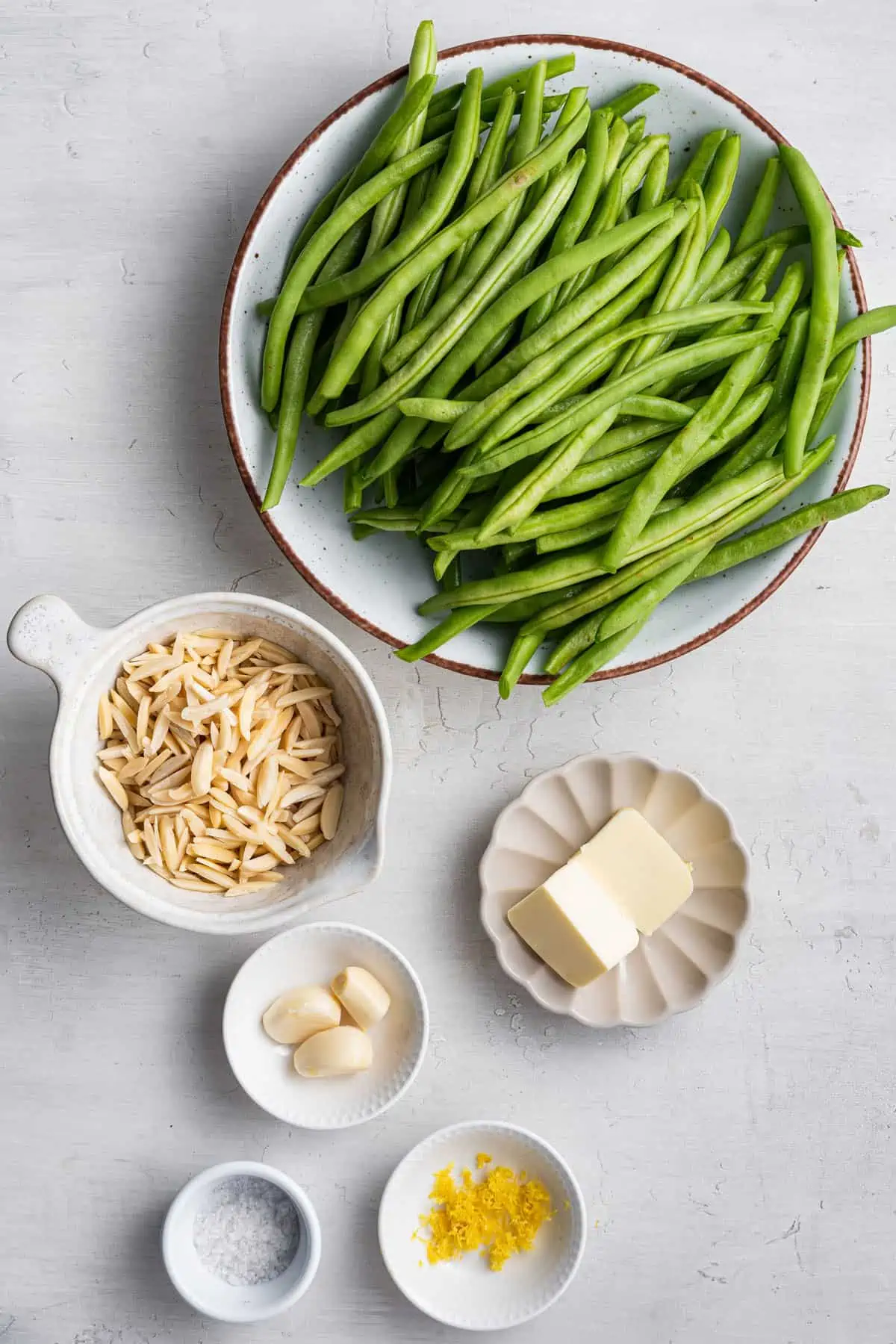 Overhead view of the ingredients for green bean almondine: a bowl of green beans, a bowl of slivered almonds, a bowl of vegan butter, a bowl of garlic, a bowl of lemon zest, and a bowl of salt
