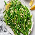 A serving tray of green beans with slivered almonds, with a lemon slice, next to a kitchen towel and two lemon slices