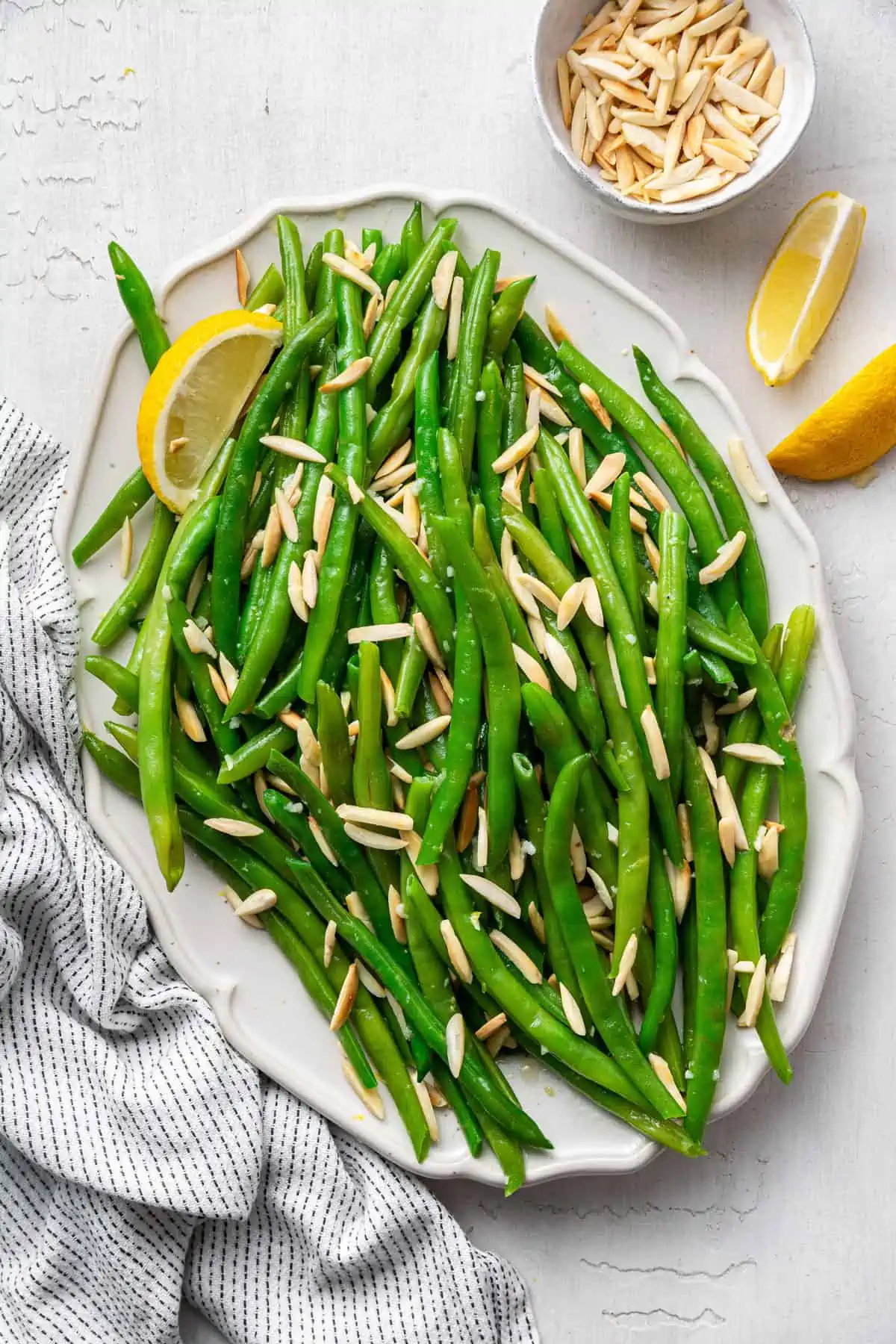 A serving tray of green beans with slivered almonds, with a lemon slice, next to a kitchen towel and two lemon slices