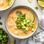 Overhead view of a bowl of white chicken chili topped with limi, chilis, avocado, and cilantro, next to another bowl of chili, a bowl of cilantro, and lime wedges