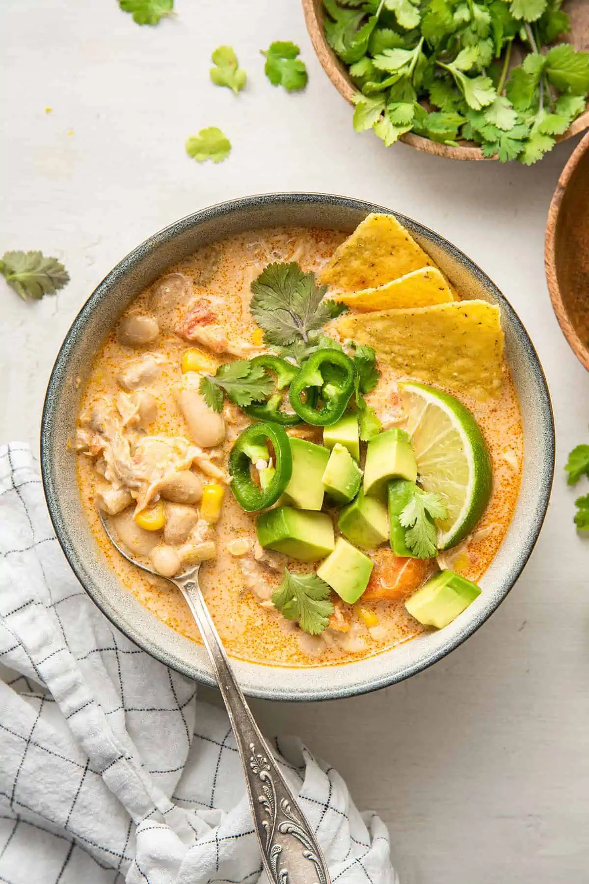 Overhead view of a bowl of white chicken chili topped with chilis, lime, avocado, cilantro, and tortilla chips, with a bowl of cilantro in the back