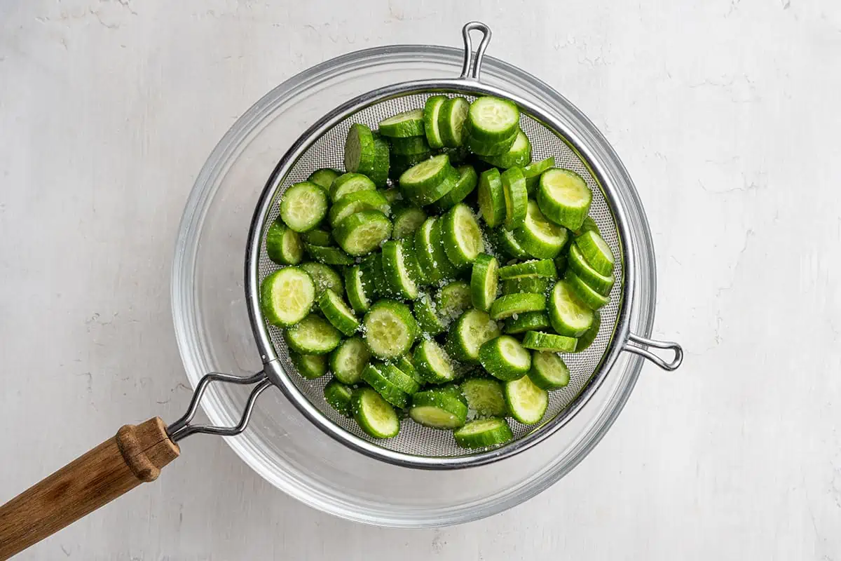 Sliced cucumbers covered in kosher salt, in a sieve over a mixing bowl