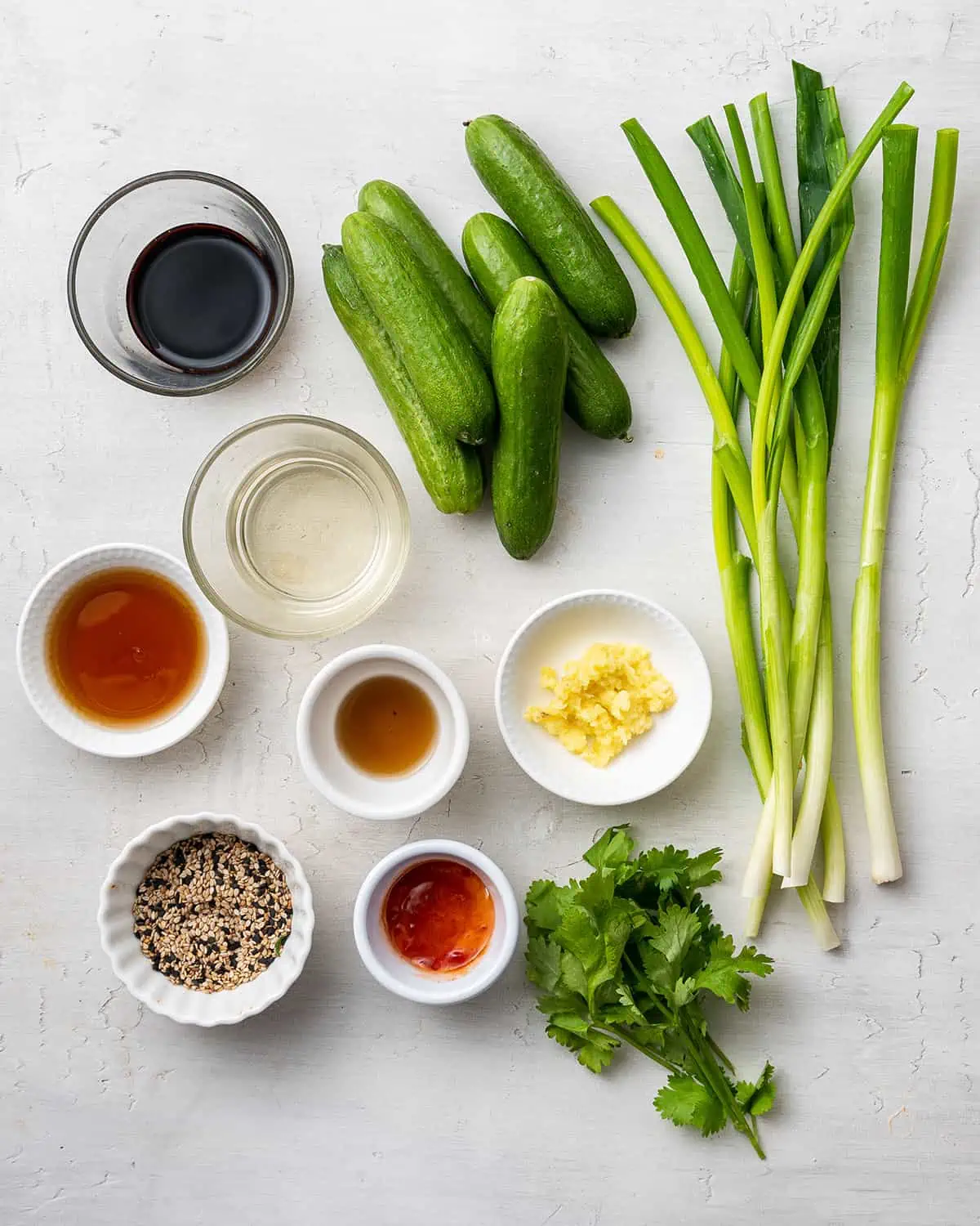 Overhead view of the ingredients needed for Asian cucumber salad: six small cucumbers, six scallions, a bunch of cilantro, and bowls of grated ginger, sesame seeds, chili garlic sauce, mirin, soy sauce, sesame oil, and fish sauce.
