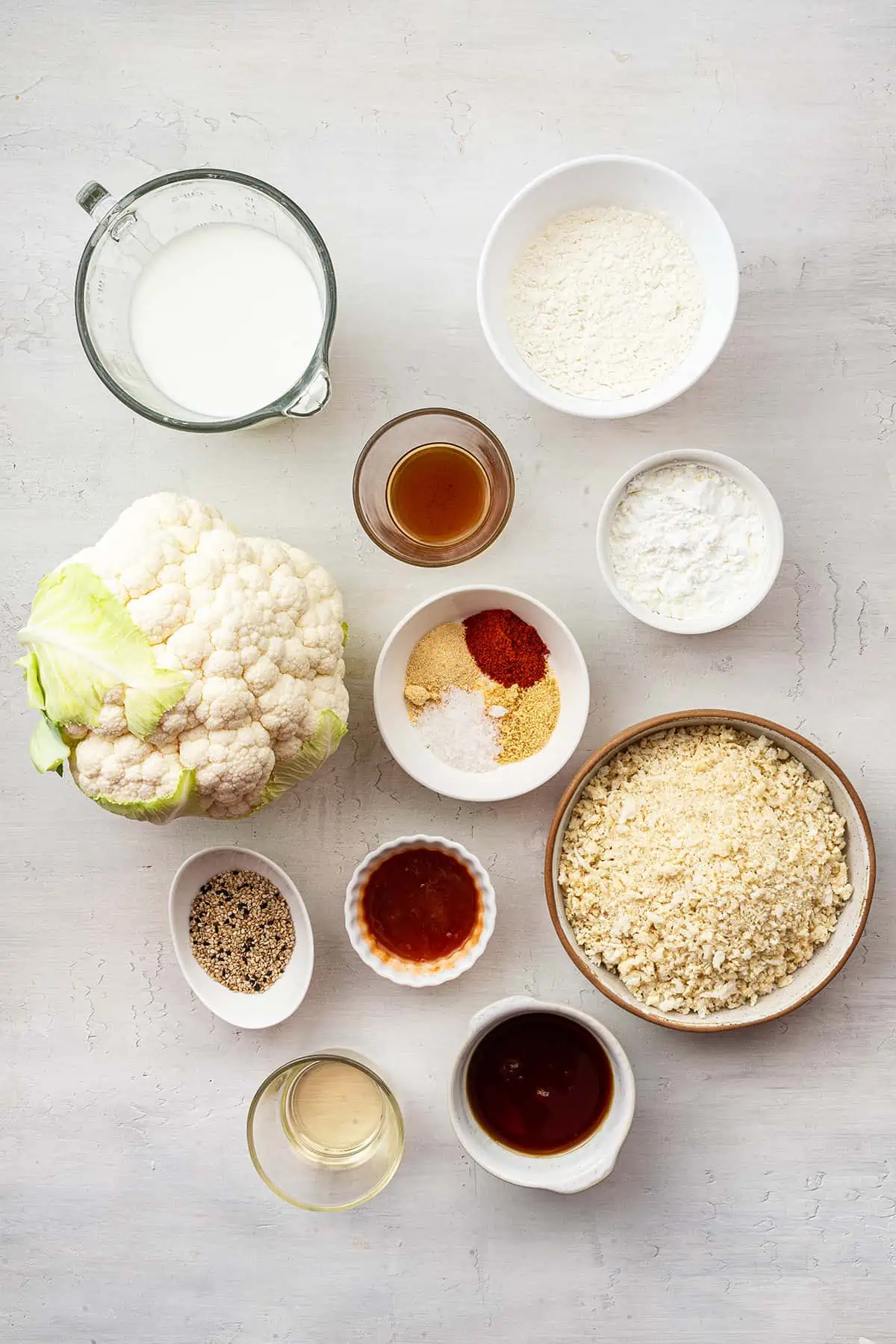 Overhead view of the ingredients for bang bang cauliflower: a head of cauliflower, a pyrex of almond milk, a bowl of seasonings, a bowl of panko bread crumbs, a bowl of chili garlic sauce, a bowl of gluten-free flour, a bowl of sesame seeds, a bowl of rice vinegar, a bowl of apple cider vinegar, a bowl of maple syrup, and a bowl of starch.