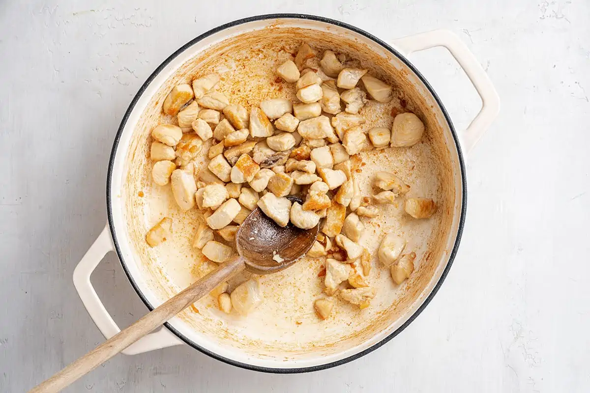 A dutch oven with chunks of chicken cooking in it, and a wooden spoon