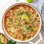 Overhead view of a pot of chicken pad thai garnished with peanuts, cilantro, and lime wedges, next to a bowl of lime wedges, a bunch of cilantro, a whole lime, and a kitchen towel