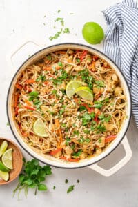 Overhead view of a pot of chicken pad thai garnished with peanuts, cilantro, and lime wedges, next to a bowl of lime wedges, a bunch of cilantro, a whole lime, and a kitchen towel