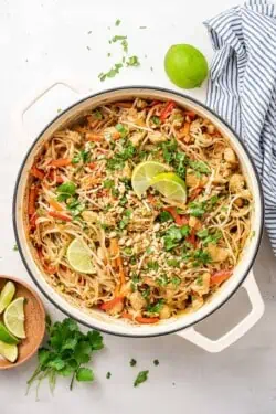 Overhead view of a pot of chicken pad thai garnished with peanuts, cilantro, and lime wedges, next to a bowl of lime wedges, a bunch of cilantro, a whole lime, and a kitchen towel