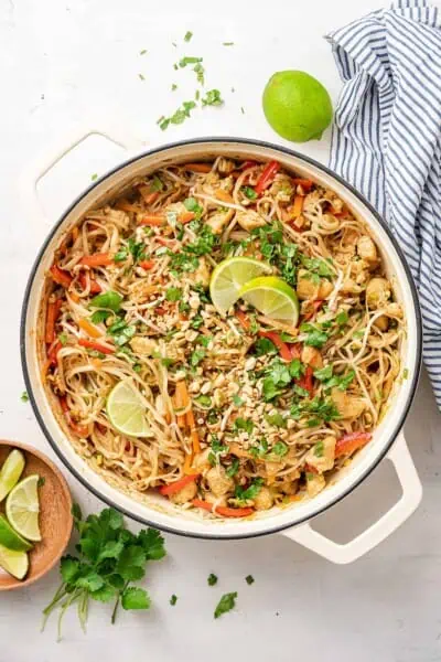Overhead view of a pot of chicken pad thai garnished with peanuts, cilantro, and lime wedges, next to a bowl of lime wedges, a bunch of cilantro, a whole lime, and a kitchen towel