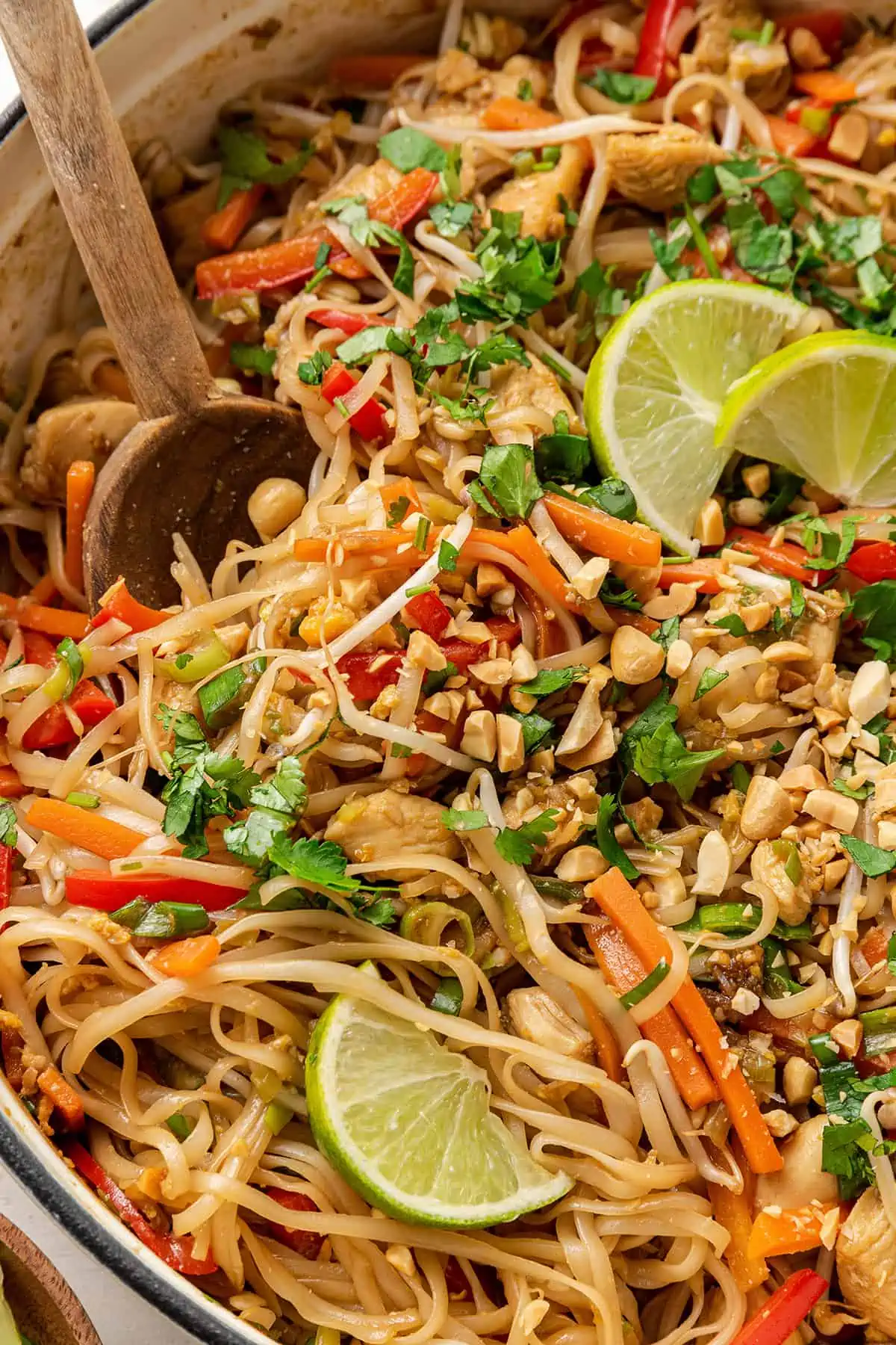 Close up of a pot of chicken pad thai garnished with cilantro, chopped peanuts, and lime wedges, with a wooden serving spoon in it