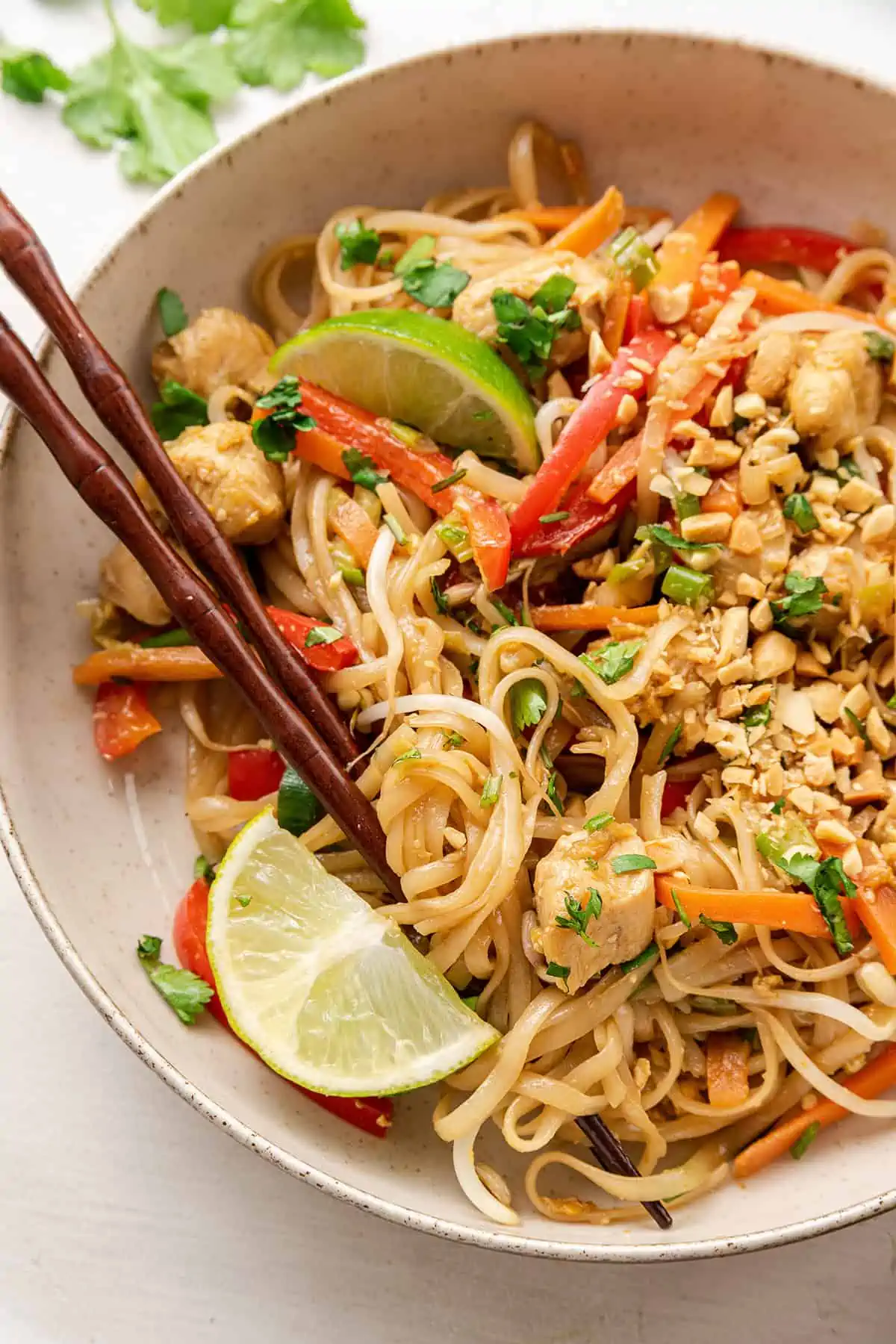 Close up of a bowl of chicken pad thai garnished with lime wedges, with two chopsticks in it
