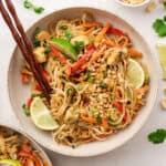 Overhead view of a bowl of chicken pad tai with lime wedges and chopsticks on top, next to a bowl of peanuts, and some lime slices and cilantro