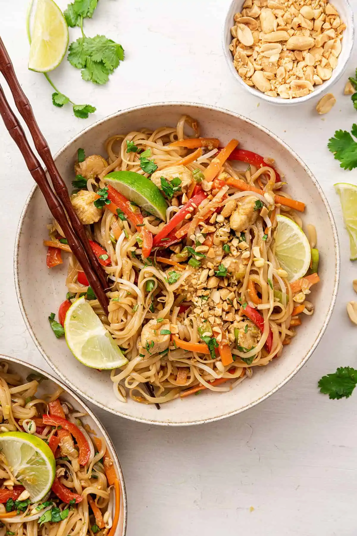 Overhead view of a bowl of chicken pad tai with lime wedges and chopsticks on top, next to a bowl of peanuts, and some lime slices and cilantro