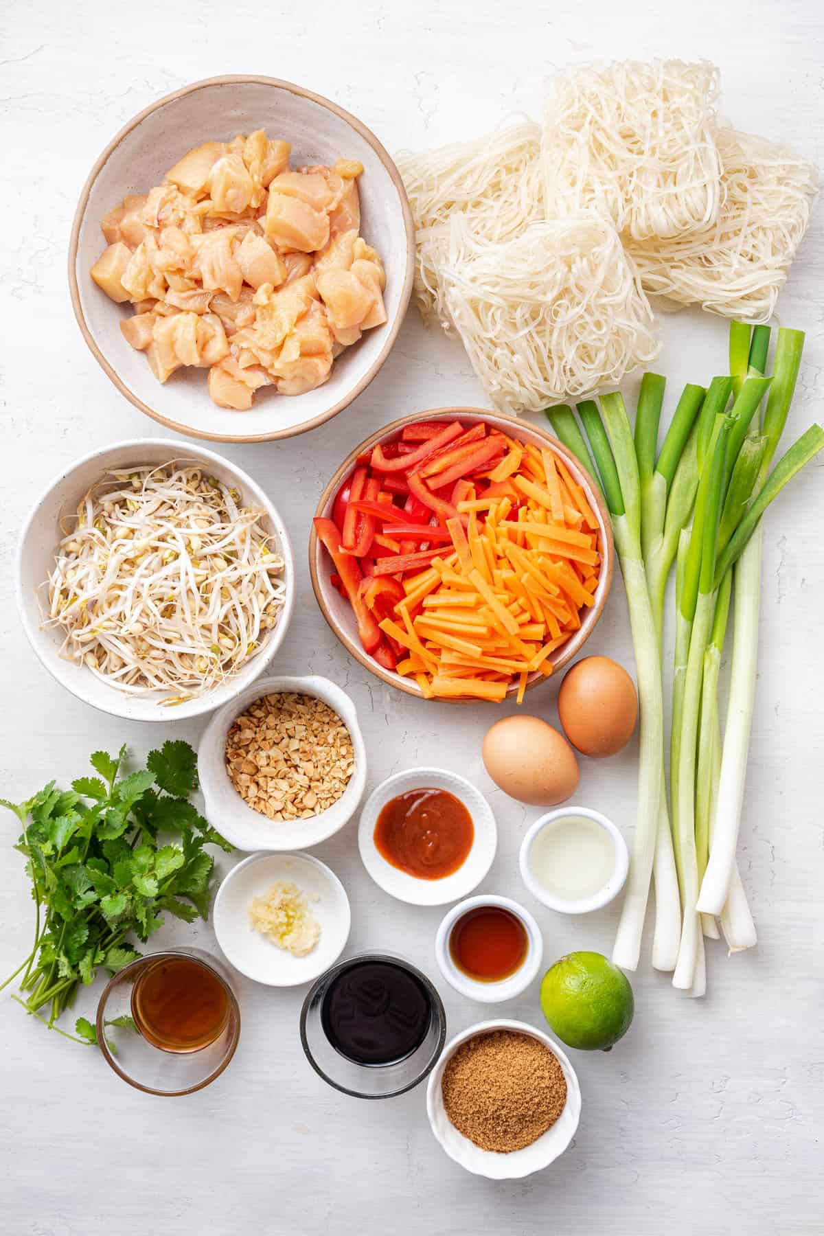 Overhead view of the ingredients needed for chicken pad thai: a bowl of cubed chicken breasts, a bowl of bean sprouts, a bowl of sliced carrots and red bell peppers, four blocks of pad thai noodles, some green onions, two eggs, a bowl of chopped peanuts, a bowl of sriracha, a bowl of chopped garlic, a bowl of soy sauce, a bowl of fish sauce, a bowl of sesame oil, a bowl of vinegar, a bowl of coconut sugar, two eggs, a lime, and a bunch of cilantro