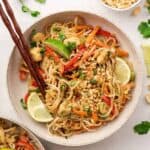 Overhead view of a bowl of chicken pad tai with lime wedges and chopsticks on top, next to a bowl of peanuts, and some lime slices and cilantro