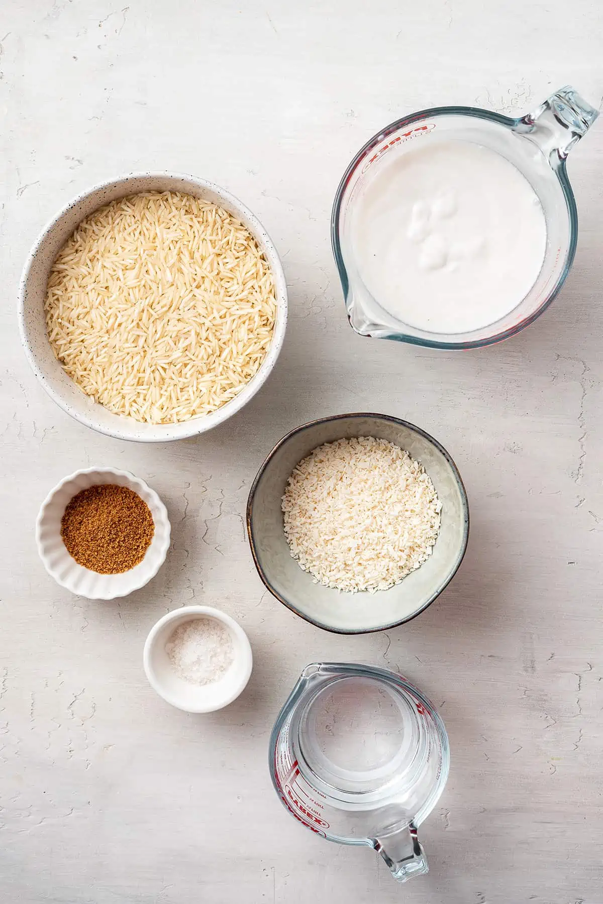 Overhead view of the ingredients needed for coconut rice: bowls of rice, shredded coconut, salt, and coconut sugar, and pyrex containers of water and coconut milk.