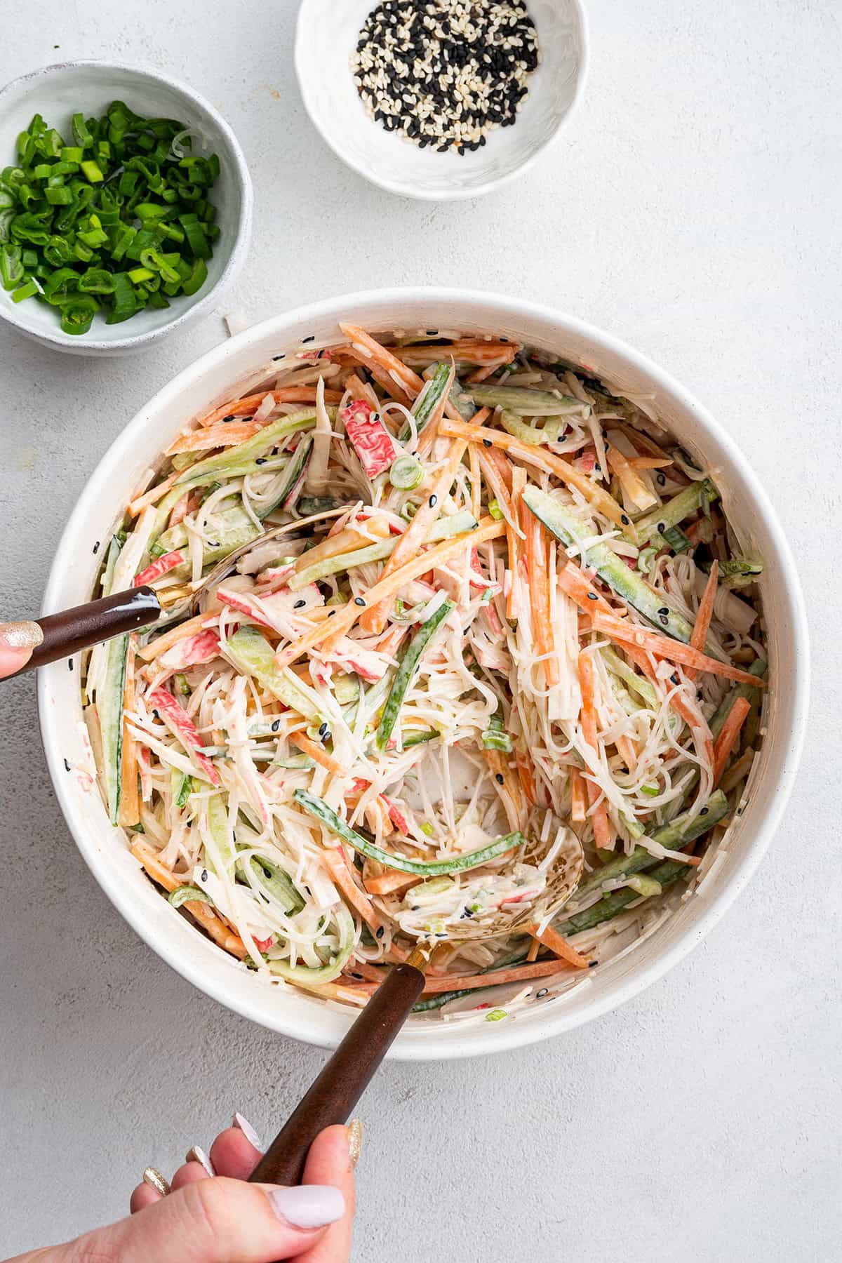 Two hands using salad spoons to toss a kani salad next to a bowl of scallions and a bowl of sesame seeds