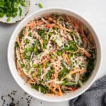 Overhead view of a bowl of kani salad topped with scallions and sesame seeds, next to a bowl of scallions, a bowl of sesame seeds, and a kitchen towel