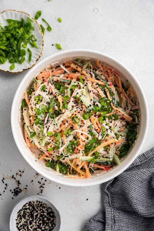 Overhead view of a bowl of kani salad topped with scallions and sesame seeds, next to a bowl of scallions, a bowl of sesame seeds, and a kitchen towel